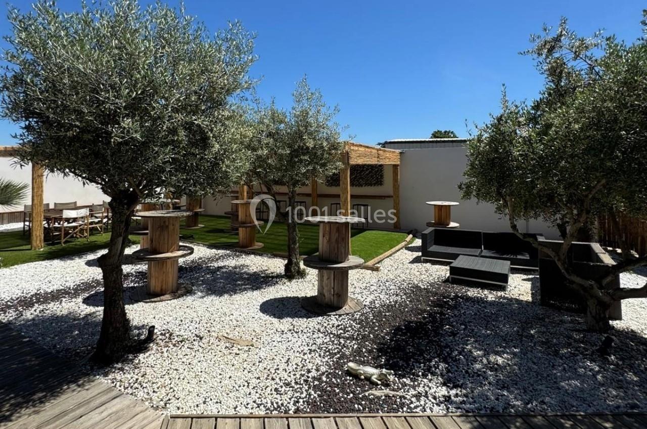 Jardin extérieur avec oliviers, gravier blanc, pelouse, tables en bois et structures en bois cylindriques.