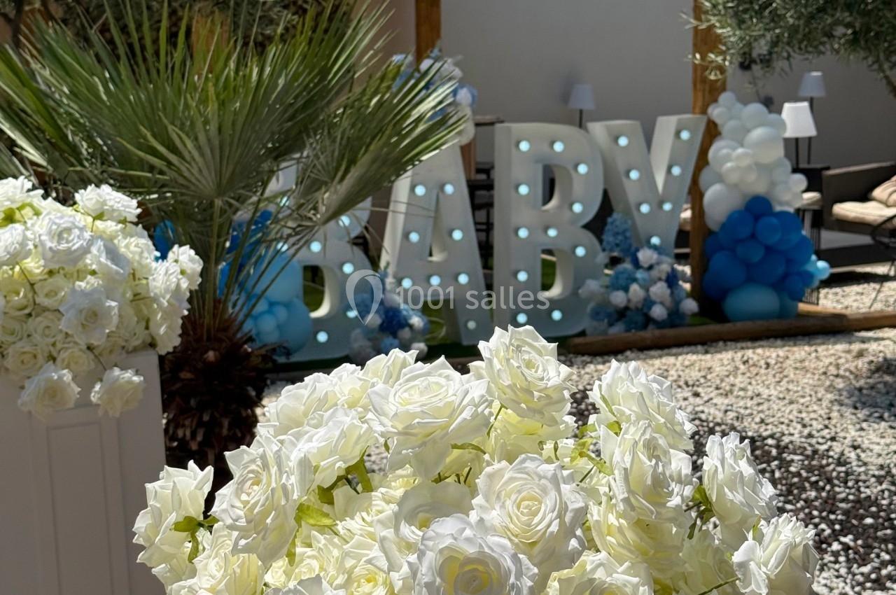 Décoration extérieure avec des roses blanches au premier plan et des ballons bleus entourant des lettres lumineuses ’BABY’.