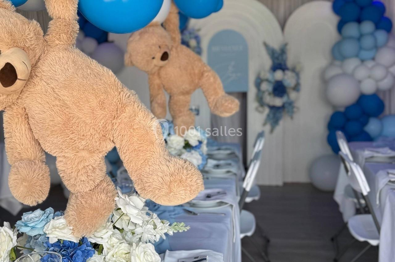 Des ours en peluche suspendus au-dessus de tables décorées de fleurs blanches et bleues, dans une salle ornée de ballons.