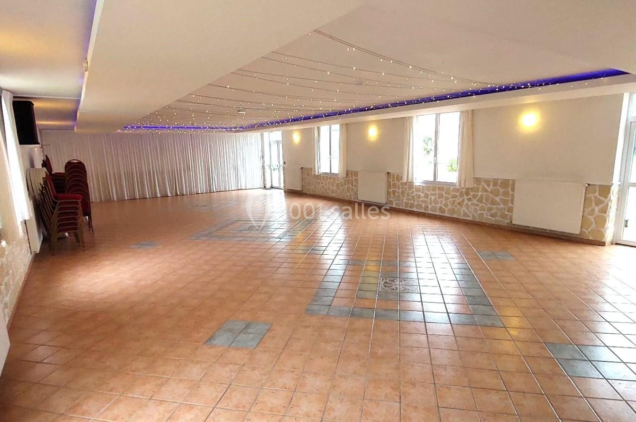 Salle lumineuse avec carrelage au sol, murs clairs, guirlandes lumineuses au plafond et chaises empilées sur le côté.