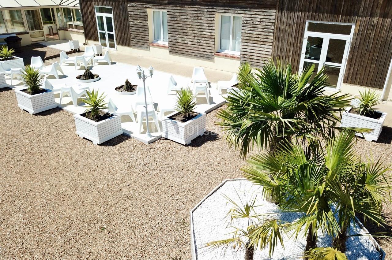 Terrasse extérieure avec mobilier blanc, plantes en pots et palmiers, devant un bâtiment en bois.