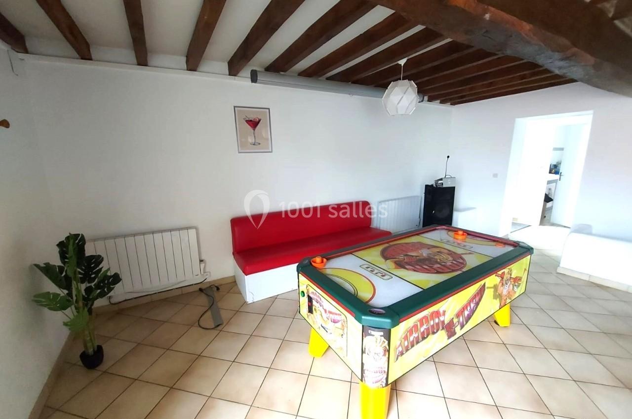 Table de air hockey colorée dans une pièce lumineuse avec canapé rouge, plante verte et poutres apparentes au plafond.