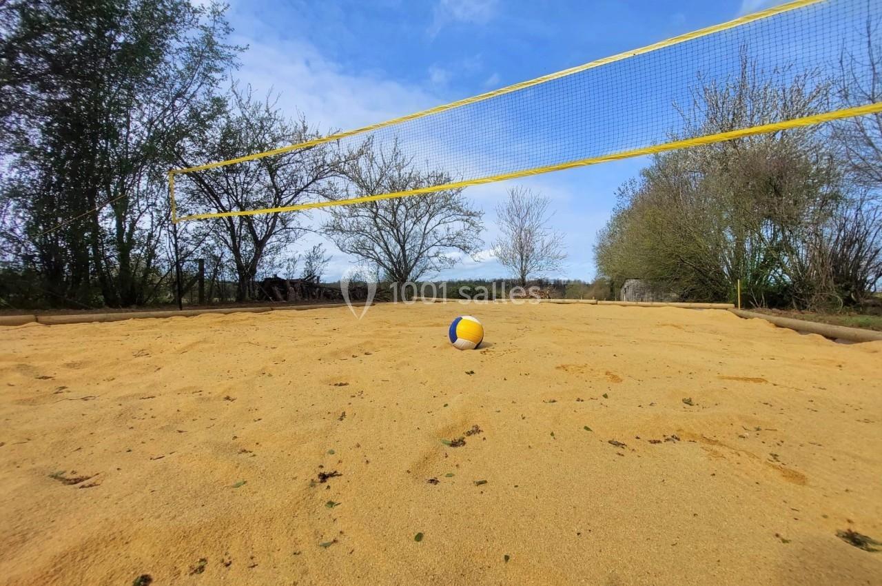Terrain de volley extérieur sur sable avec un ballon au centre, entouré d'arbres sous un ciel bleu.