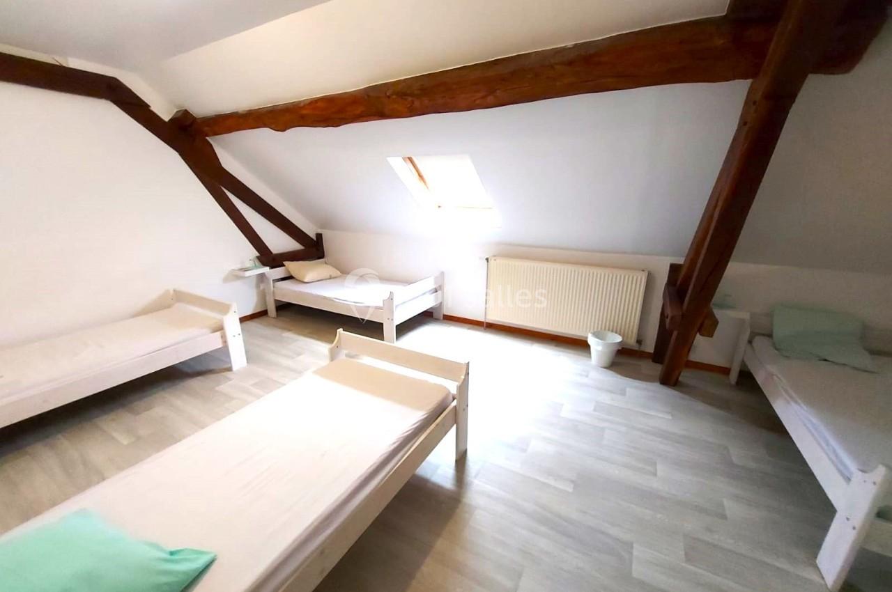 Chambre mansardée avec quatre lits simples, parquet clair, poutres apparentes et lumière naturelle provenant d'une fenêtre.