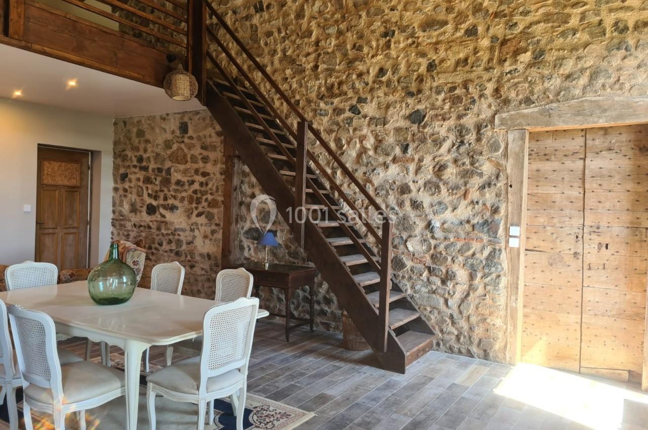 Salle à manger avec table blanche, chaises en osier, escalier en bois, murs en pierre et porte en bois massif.