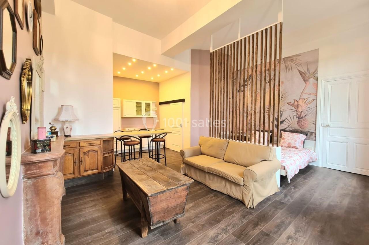 Salon lumineux avec canapé beige, table basse en bois, cuisine ouverte et séparation en bois devant un lit.