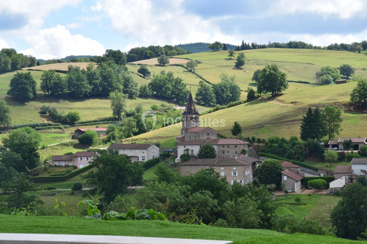 Village rural avec église au clocher pointu, entouré de collines verdoyantes et de prairies sous un ciel partiellement…