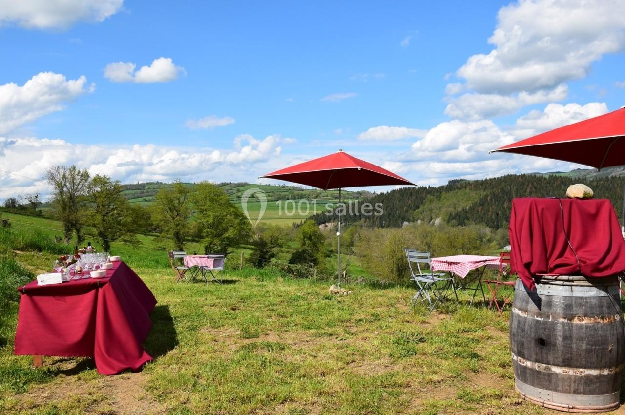 Tables et chaises installées en plein air sur une pelouse, avec nappes rouges et parasols, vue sur une campagne vallonnée.