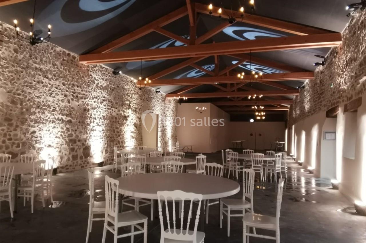 Salle avec murs en pierre apparente, poutres en bois, tables rondes et chaises blanches, éclairage tamisé.