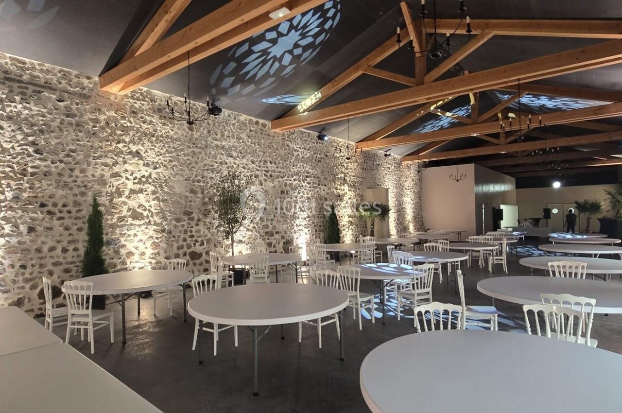 Salle de réception avec murs en pierre, poutres apparentes, tables rondes blanches et chaises alignées.
