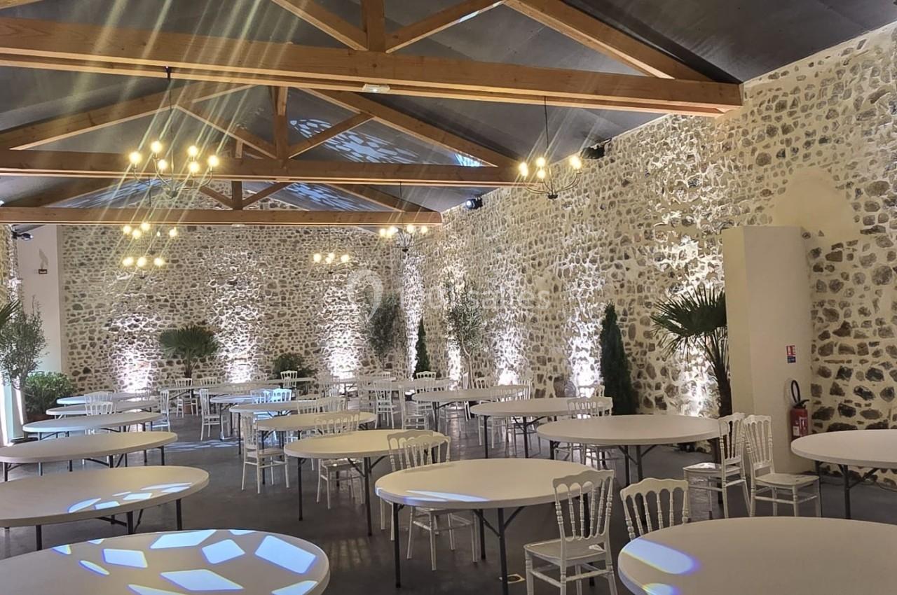 Salle en pierre avec poutres apparentes, tables rondes blanches, chaises et éclairage décoratif.