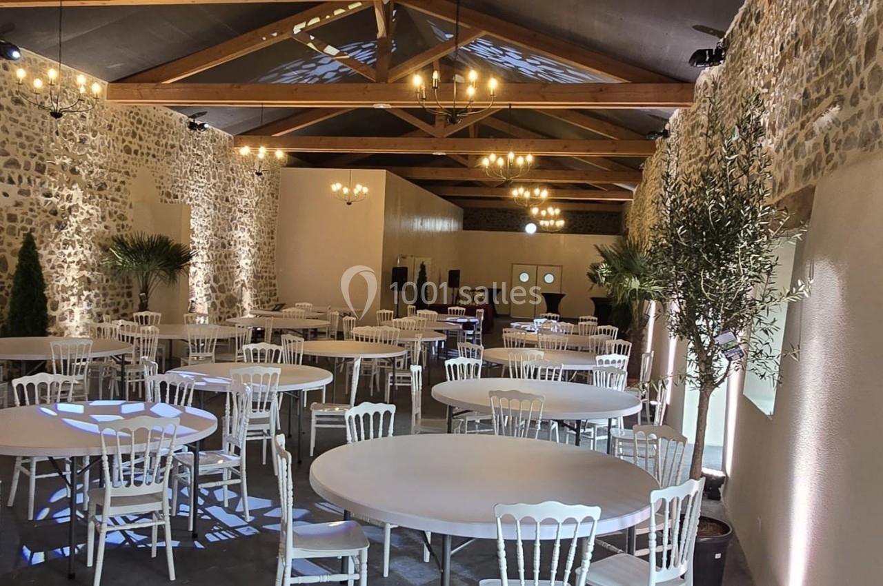 Salle en pierre avec des tables rondes et des chaises blanches, éclairée par des lustres et des spots au plafond.