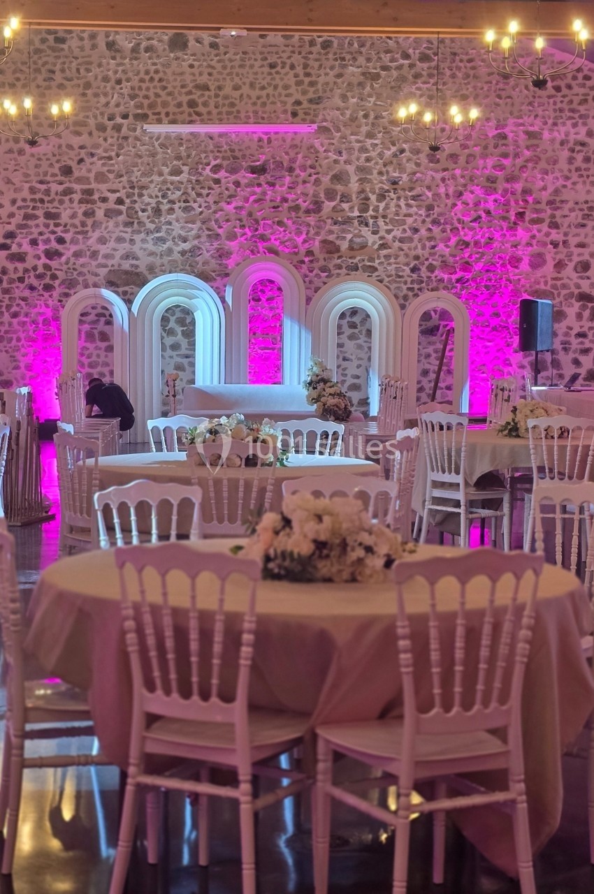 Salle de réception avec tables décorées, chaises blanches, éclairage rose et murs en pierre apparente.