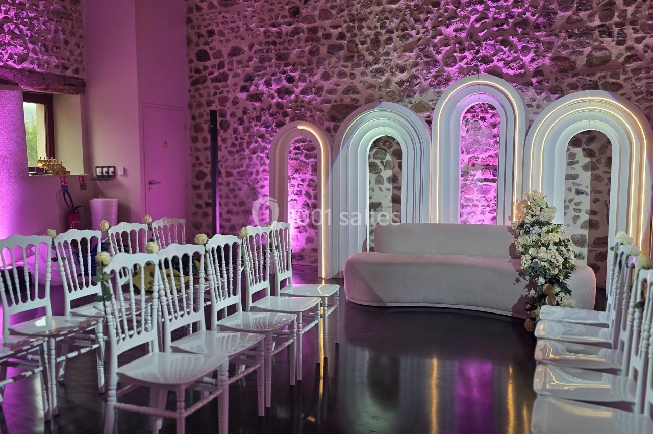 Salle décorée pour un événement, avec des chaises blanches alignées, un canapé et des arches lumineuses sur fond de murs en…