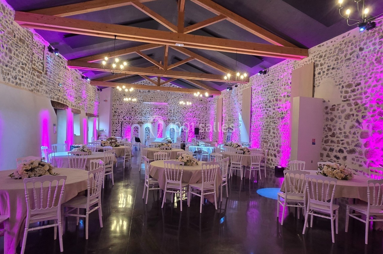 Salle de réception en pierre avec poutres apparentes, éclairage violet et tables rondes décorées de fleurs blanches.