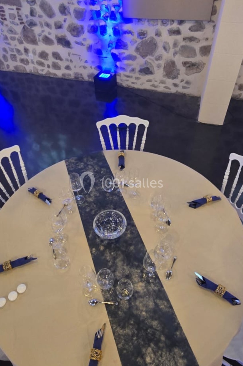 Table ronde dressée avec une nappe beige, chemin de table bleu, serviettes pliées et verres, dans une salle en pierre.