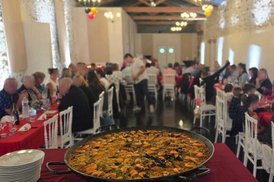 Grande paella servie dans un plat rond au premier plan, avec des convives attablés dans une salle de réception décorée.