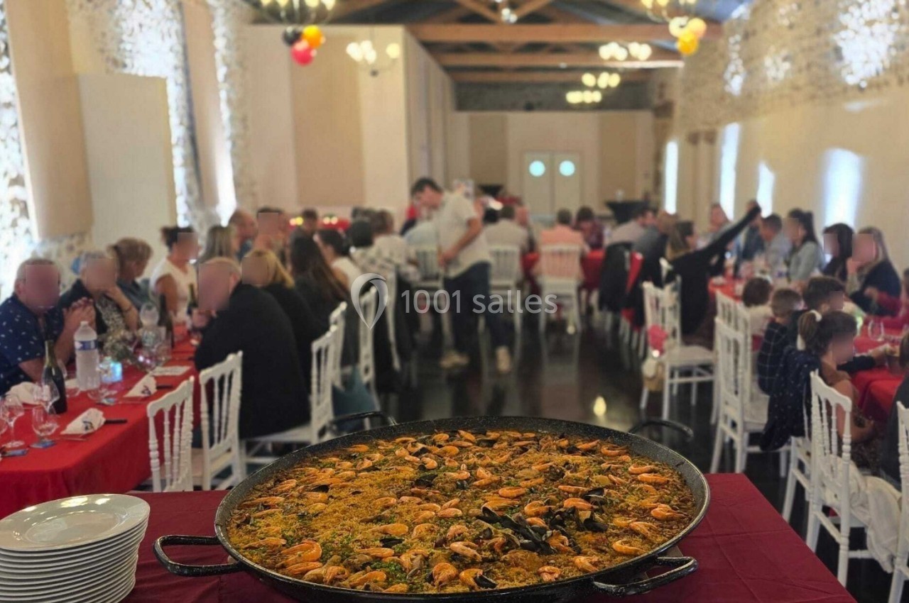 Grande paella servie dans un plat rond au premier plan, avec des convives attablés dans une salle de réception décorée.