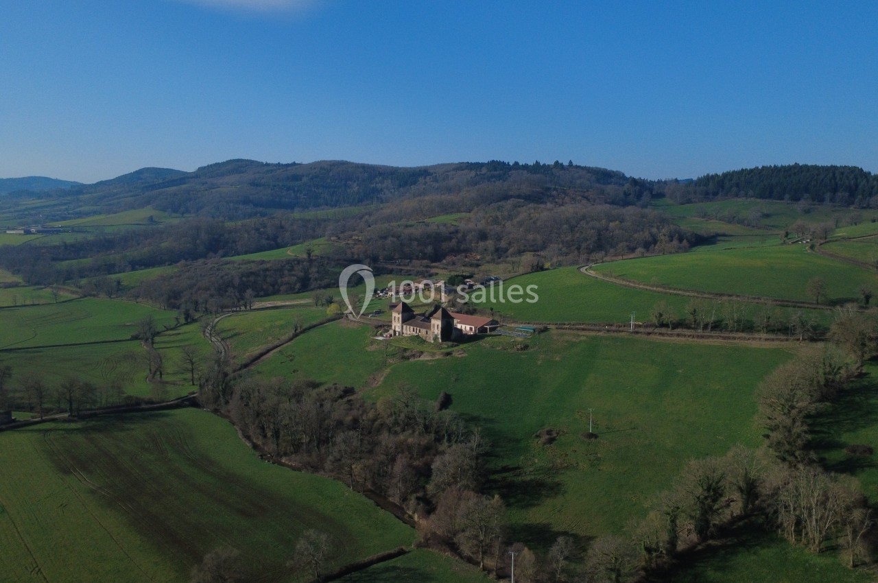 Vue aérienne d'une campagne vallonnée avec une ferme isolée entourée de champs et de forêts sous un ciel bleu.