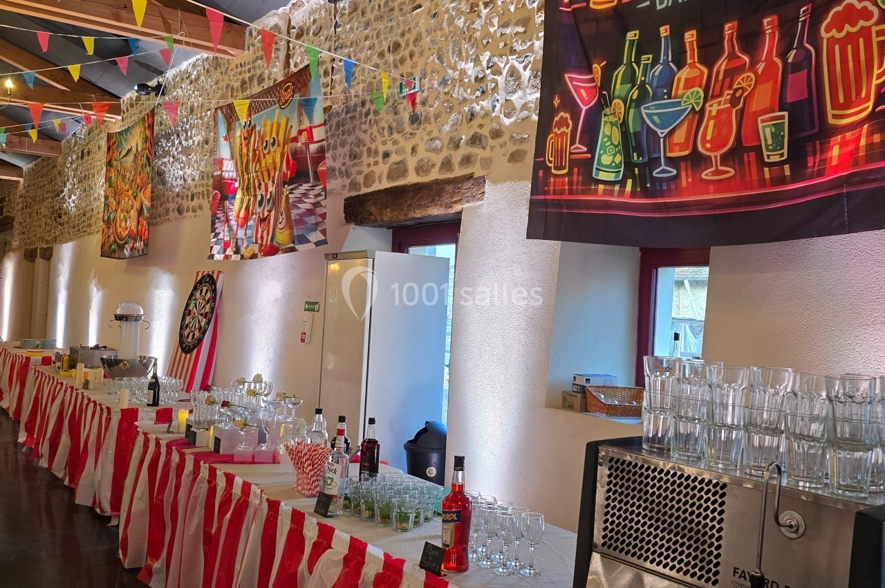 Salle décorée pour un événement festif avec tables garnies de verres, boissons et banderoles colorées accrochées aux murs.