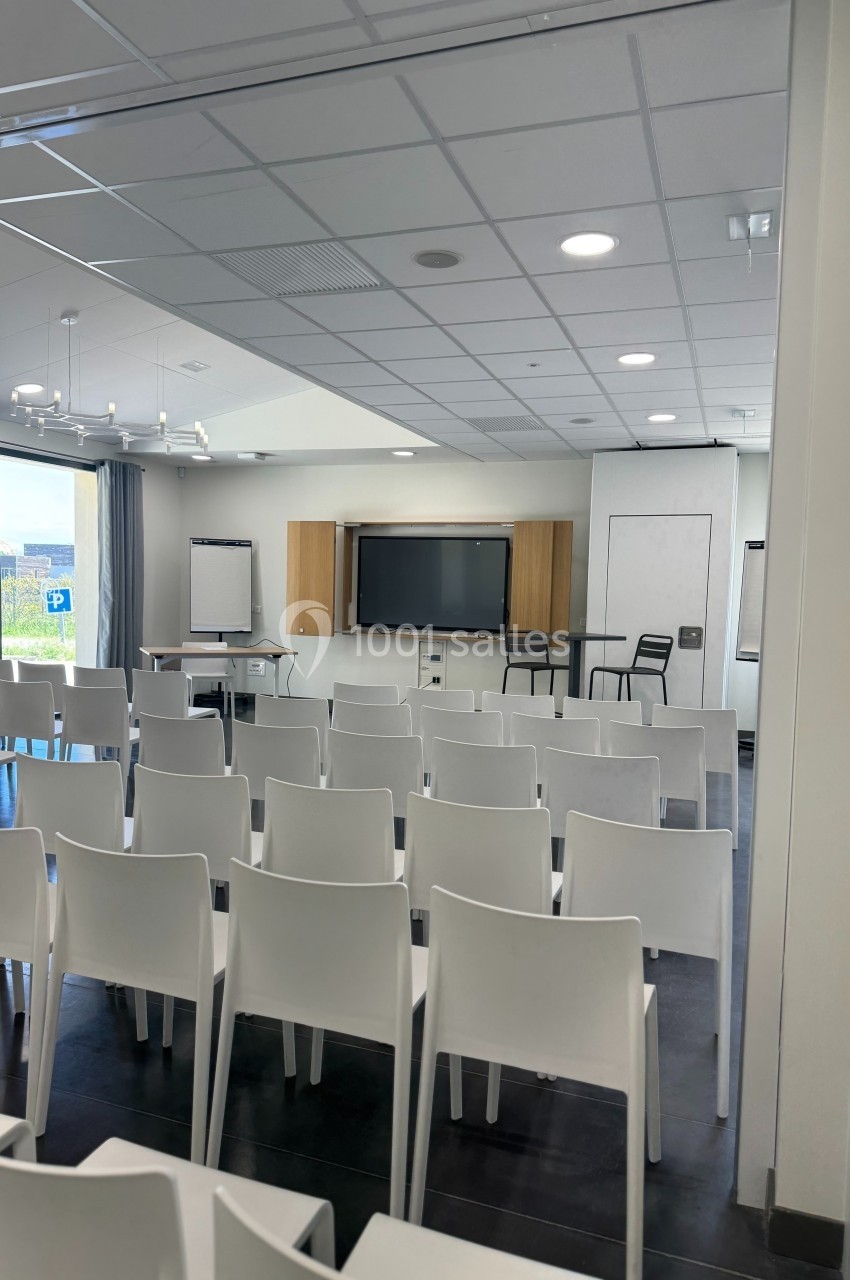Salle de conférence lumineuse avec chaises blanches alignées, tableau interactif et fenêtres donnant sur l'extérieur.