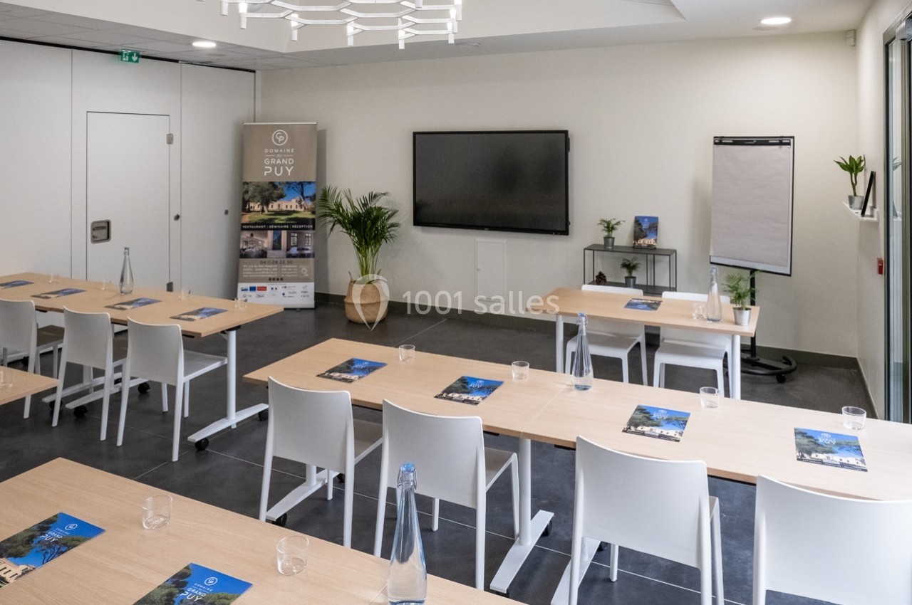 Salle de réunion lumineuse avec tables, chaises blanches, écran mural, paperboard et brochures disposées sur les tables.