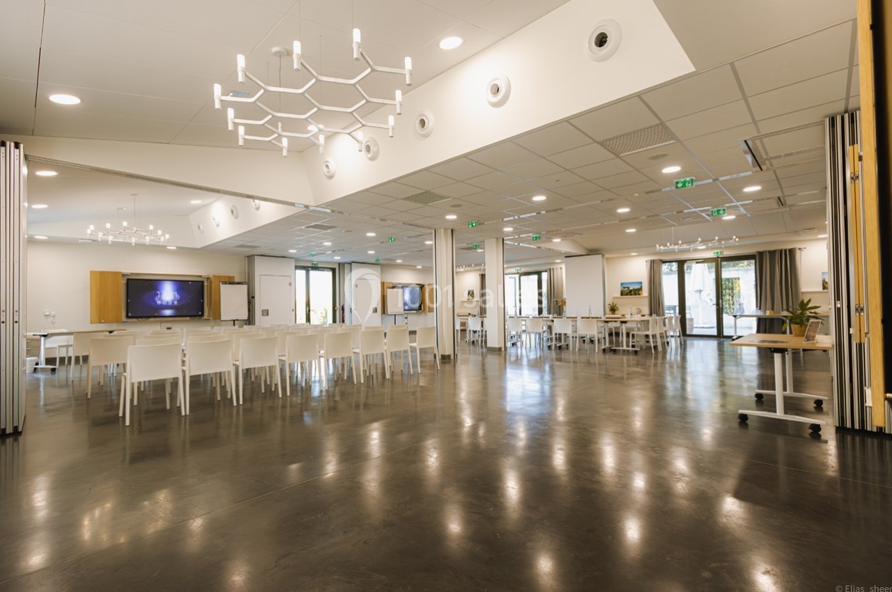 Salle de réunion spacieuse et lumineuse avec des chaises blanches, des tables et des écrans muraux.