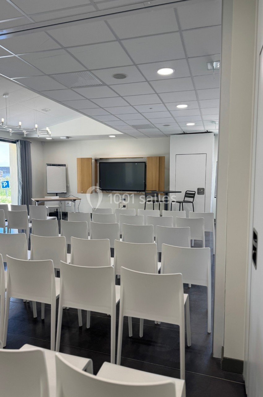 Salle de conférence lumineuse avec rangées de chaises blanches, tableau interactif et paperboard à l'avant.