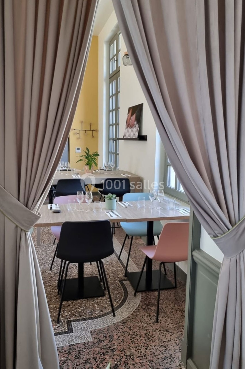Salle de restaurant lumineuse avec tables dressées, chaises colorées et rideaux ouverts encadrant la vue.