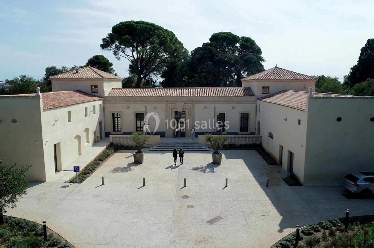 Bâtiment ancien restauré avec façade beige, cour pavée, arbres en arrière-plan et deux personnes au centre.
