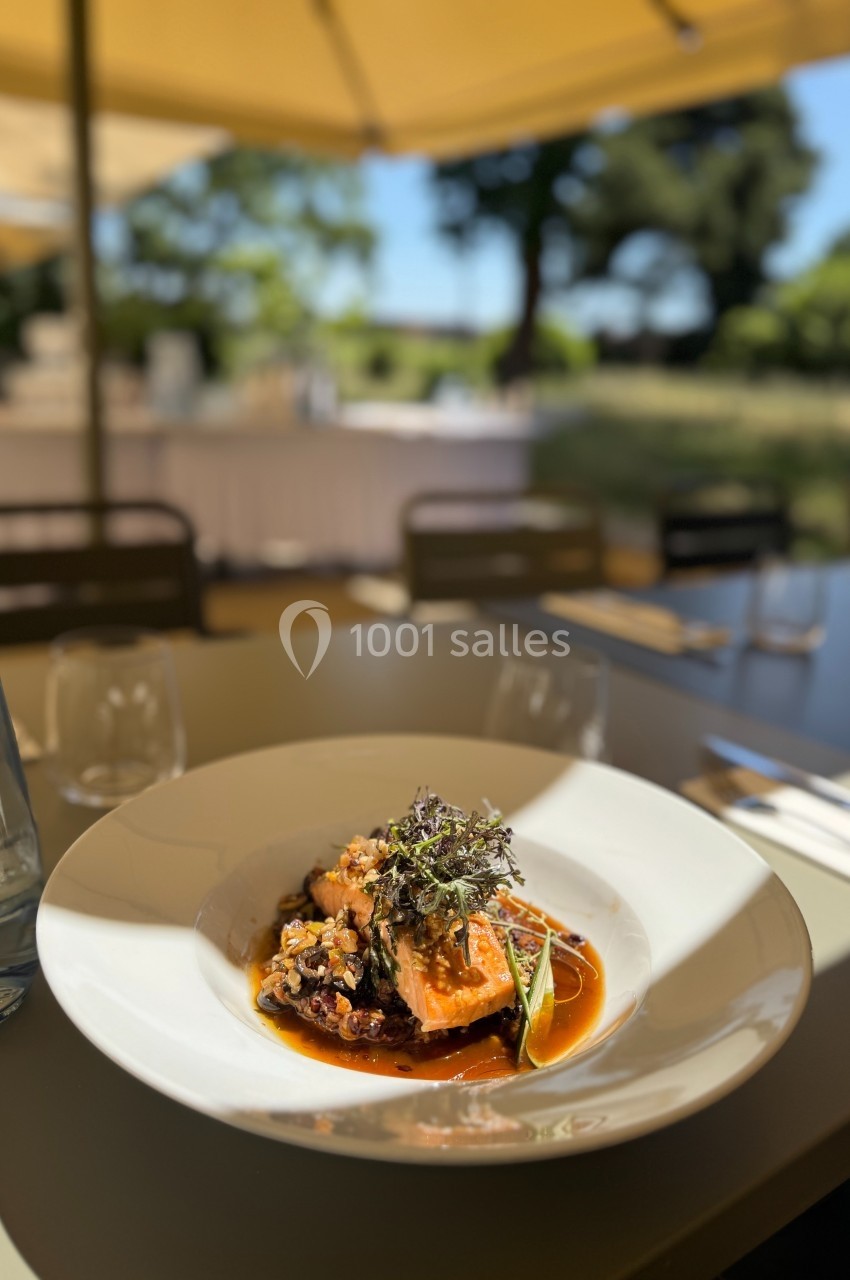 Plat gastronomique servi en terrasse, composé de légumes et viande dans une sauce, avec une table dressée en arrière-plan.