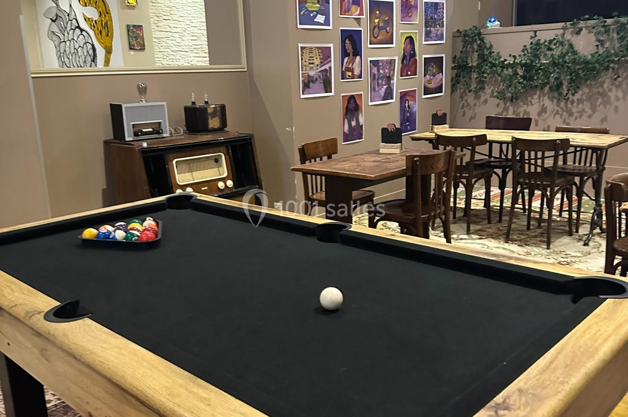 Table de billard noire dans une salle décorée de meubles en bois, affiches colorées et plantes murales.