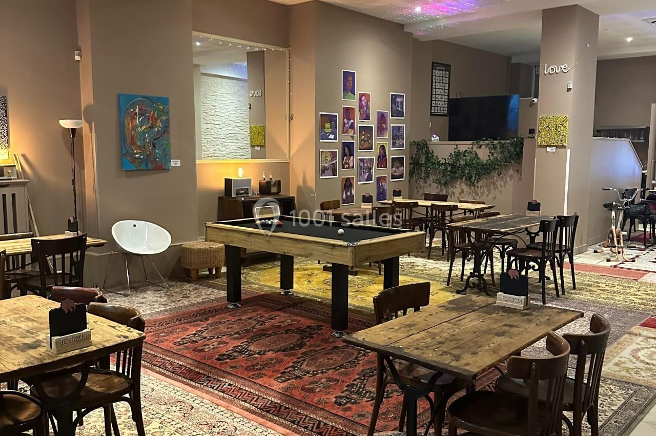 Salle conviviale avec tables en bois, tapis colorés, billard central, décorations murales et éclairage chaleureux.