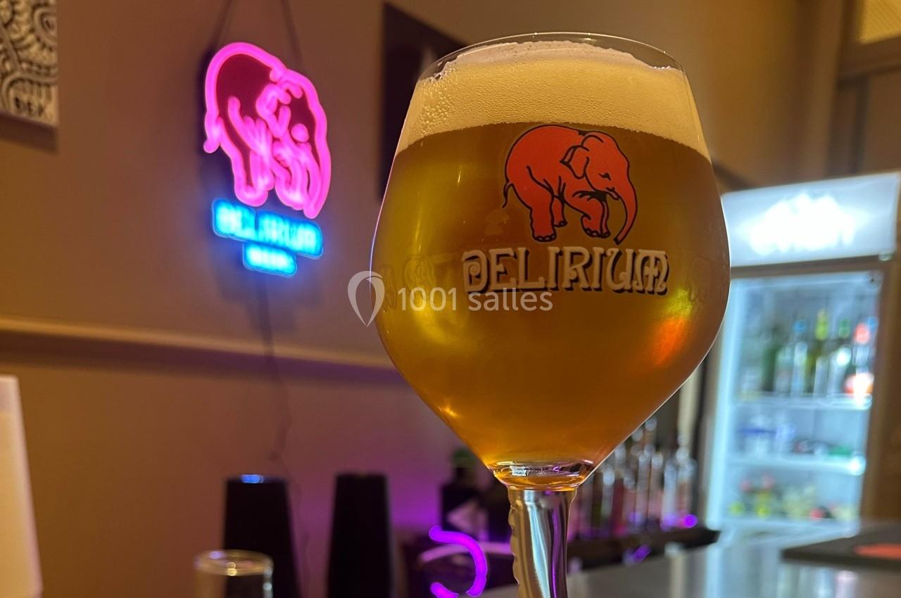 Verre de bière Delirium dorée avec mousse, posé dans un bar éclairé par une enseigne néon rose en arrière-plan.