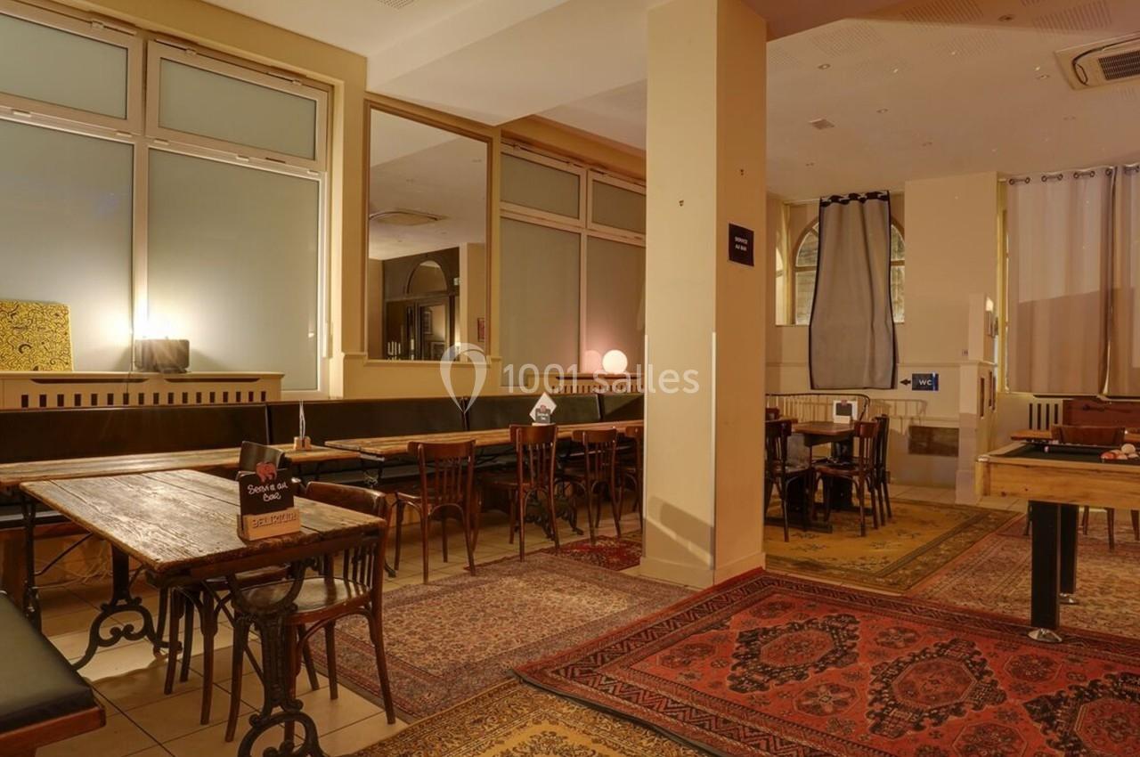Salle avec tables en bois, chaises, banquettes, tapis colorés et baby-foot dans un espace lumineux et chaleureux.