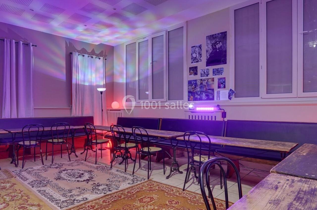 Salle avec tables en bois, chaises en métal, tapis au sol et éclairage coloré diffus sur les murs.
