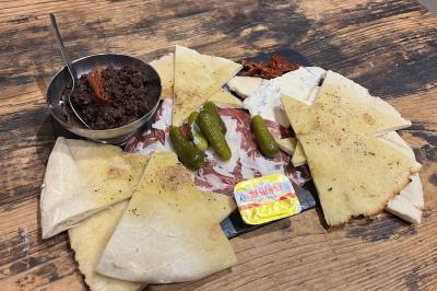 Assiette de charcuterie avec pain plat, cornichons, tapenade, fromage et beurre sur une table en bois.