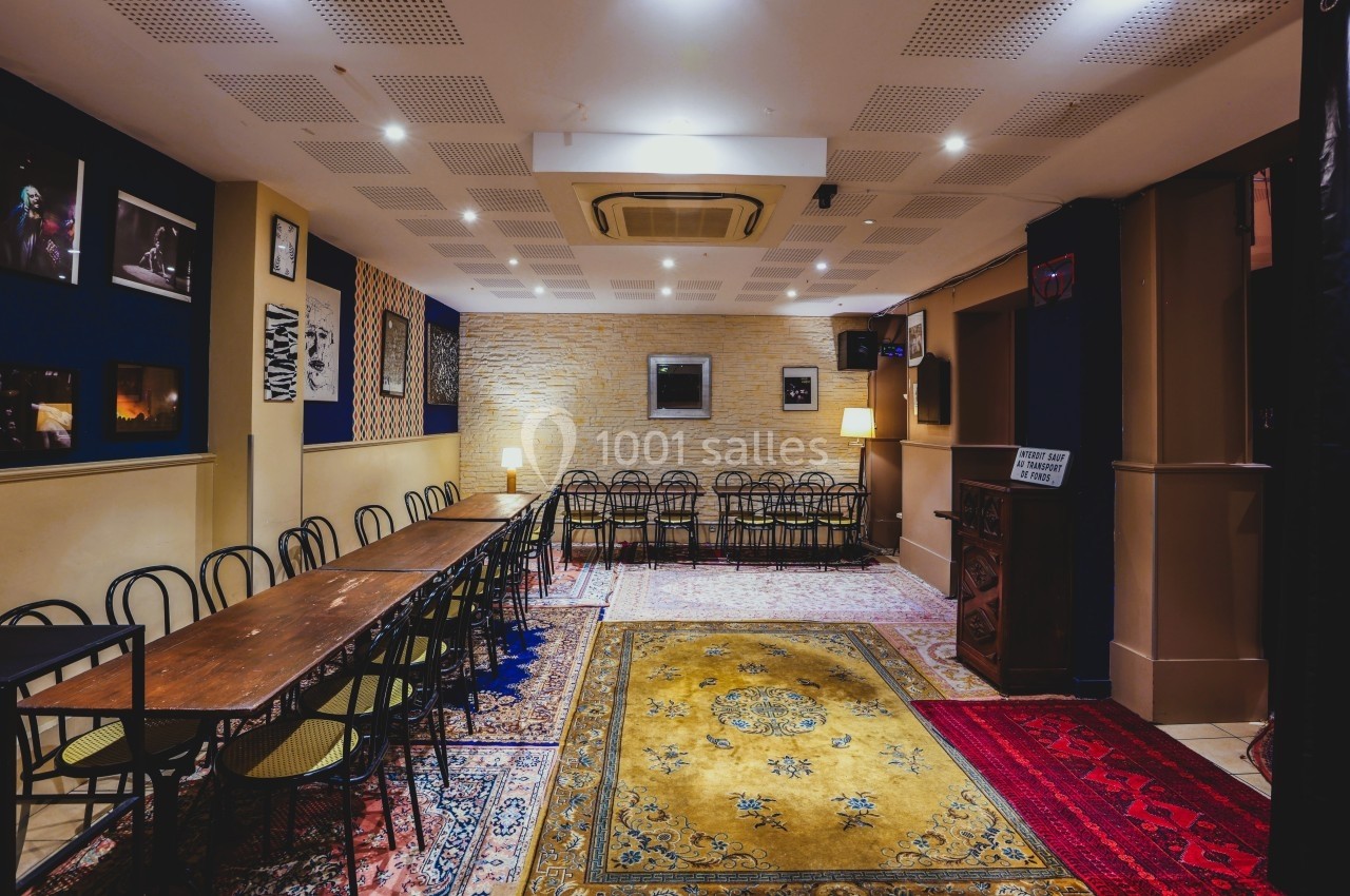 Salle lumineuse avec longues tables en bois, chaises noires, tapis colorés et murs décorés de cadres et photos.