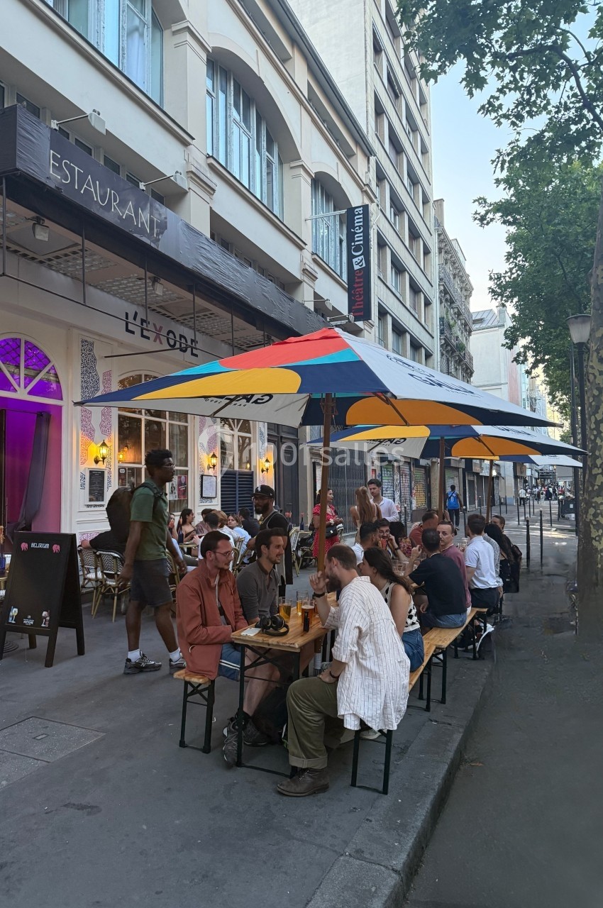 Des personnes dînent à des tables en terrasse sous des parasols colorés devant un restaurant en ville.