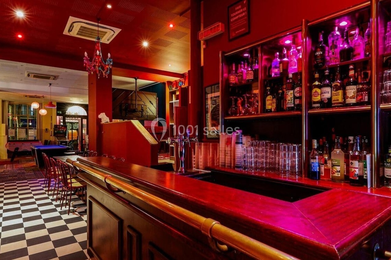 Bar avec comptoir en bois, étagères remplies de bouteilles, éclairage rouge et sol en damier noir et blanc.