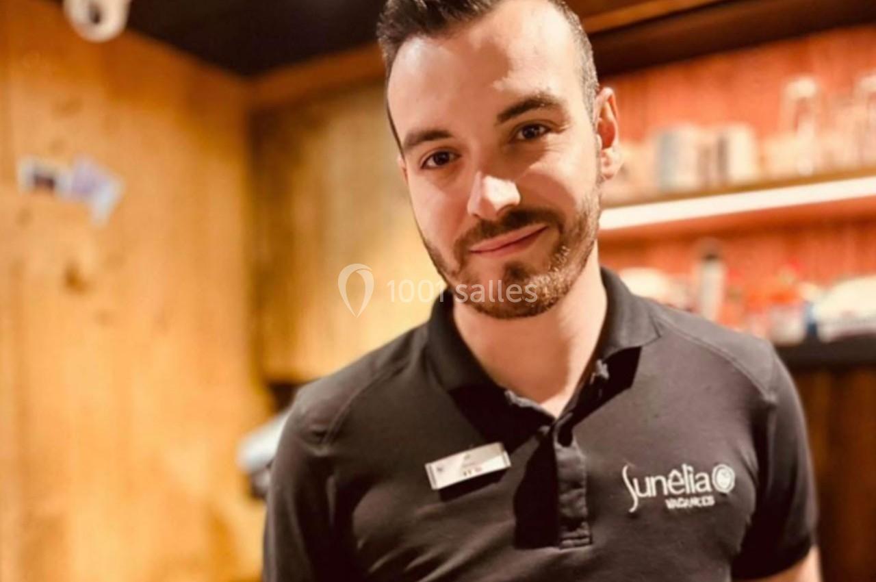 Homme souriant portant un polo noir avec un logo, dans un intérieur en bois chaleureux.