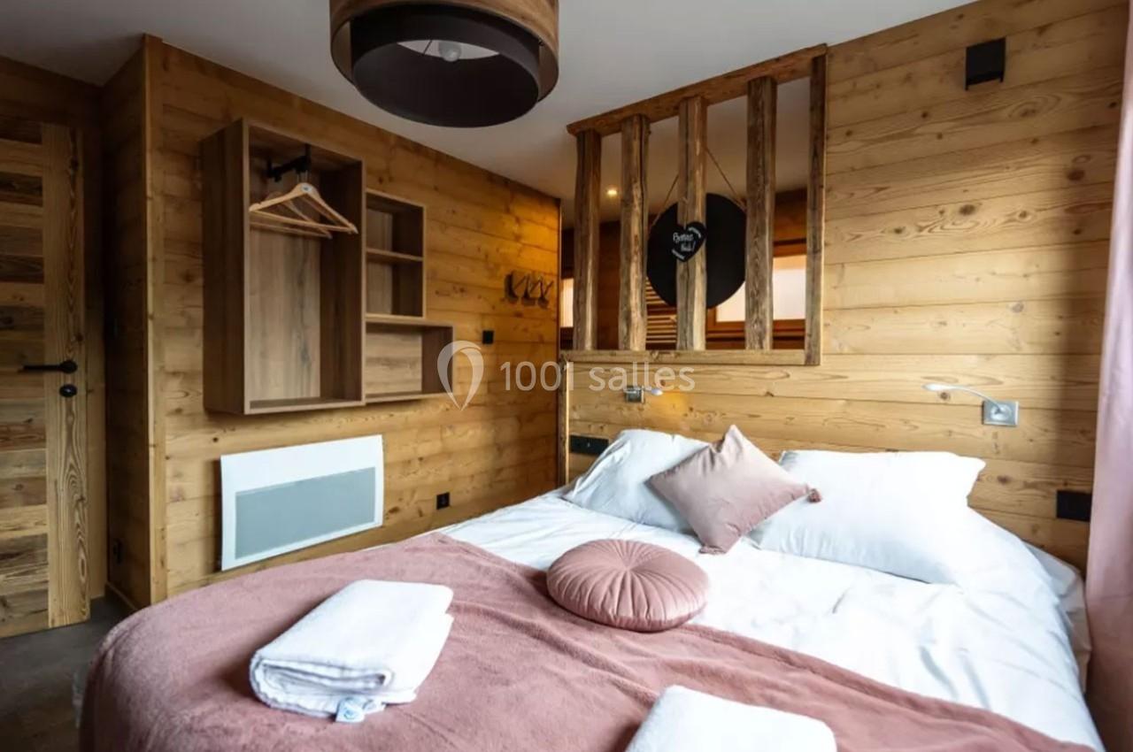 Chambre en bois avec lit double, coussins roses, serviettes pliées et penderie ouverte dans un décor chaleureux.