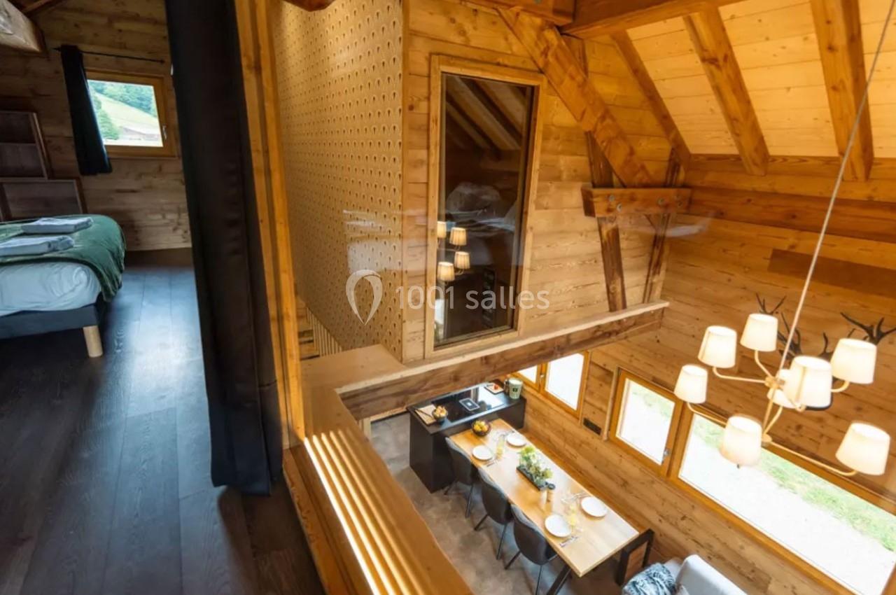 Vue intérieure d'un chalet en bois avec une mezzanine, une salle à manger lumineuse et une chambre adjacente.
