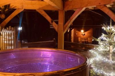 Bain à remous éclairé en violet sous une pergola en bois, avec un sapin décoré et un chalet en arrière-plan.