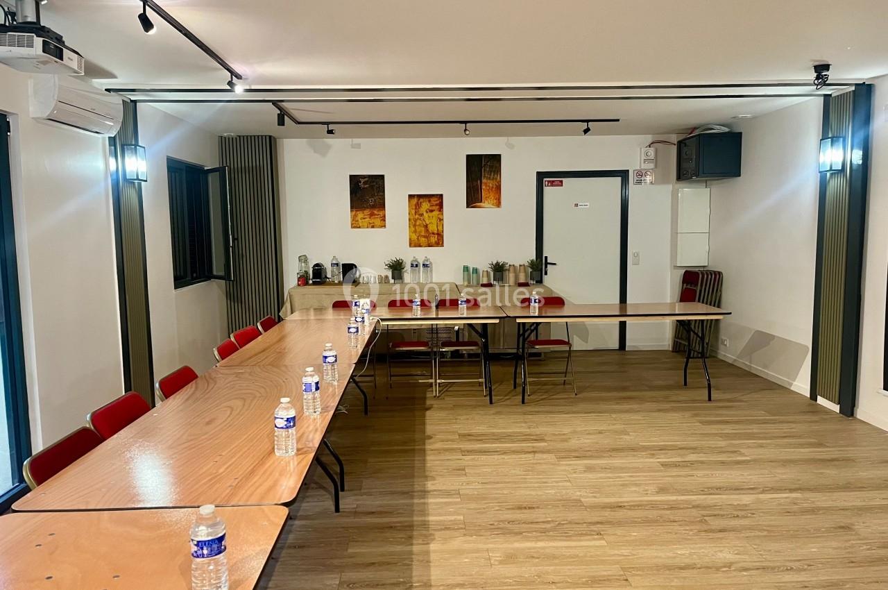 Salle de réunion avec tables en bois disposées en U, chaises rouges, bouteilles d'eau et coin café en arrière-plan.