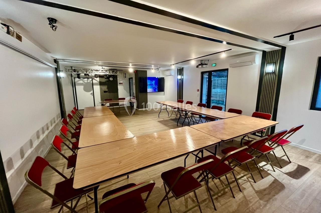 Salle de réunion lumineuse avec tables en bois disposées en U et chaises rouges, écran au mur en arrière-plan.
