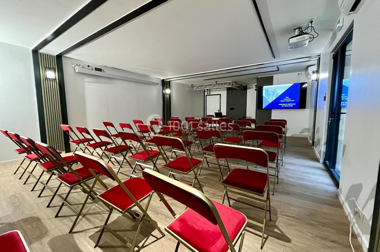 Salle de conférence avec des chaises rouges alignées face à un écran de projection et un tableau blanc.
