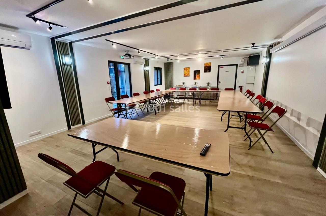 Salle de réunion lumineuse avec tables disposées en U, chaises rouges et équipement audiovisuel.
