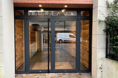 Entrée d'un immeuble avec porte vitrée, numéro 16 et inscription ’Bureaux’, sol pavé avec quelques feuilles.
