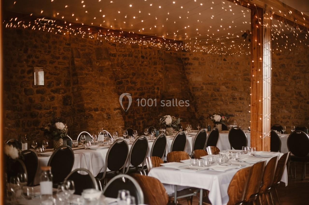 Salle de réception décorée avec des guirlandes lumineuses, des tables dressées et des murs en pierre apparente.